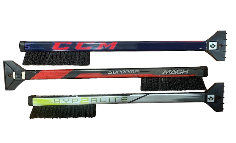 May include: Three hockey stick ice scrapers with black bristles. The top scraper is blue with the word "CCM" printed on it. The middle scraper is red with the words "Supreme" and "MACH" printed on it. The bottom scraper is gray with the words "HYPERLITE" printed on it.