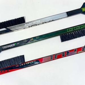 May include: Three hockey stick ice scrapers with brushes. The top scraper is silver with black and white Bauer branding. The middle scraper is green with black and white CCM branding. The bottom scraper is red with black and white TRUE branding.