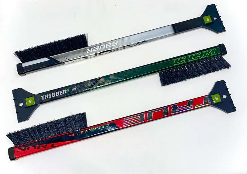 May include: Three hockey stick ice scrapers with brushes. The top scraper is silver with black and white Bauer branding. The middle scraper is green with black and white CCM branding. The bottom scraper is red with black and white TRUE branding.
