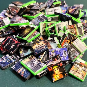 May include: A pile of miniature video game cases, many with green borders, featuring various game titles and colorful cover art. The cases are stacked and scattered, showcasing a collection of PlayStation 2 and other console games.