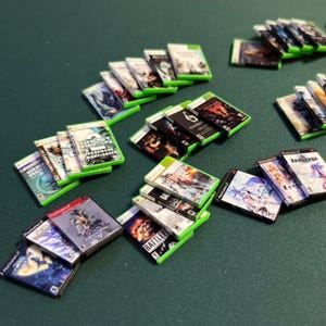 May include: A collection of miniature video game cases, including titles like "Ghost Recon" and "Battlefield", are arranged on a green surface. The cases feature colorful artwork and text, with green and black cases. Some are labeled "Playstation 2".