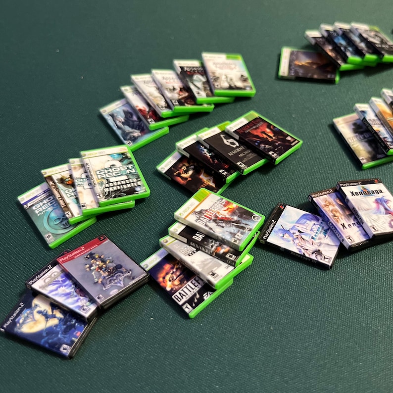 May include: A collection of miniature video game cases, including titles like "Ghost Recon" and "Battlefield", are arranged on a green surface. The cases feature colorful artwork and text, with green and black cases. Some are labeled "Playstation 2".