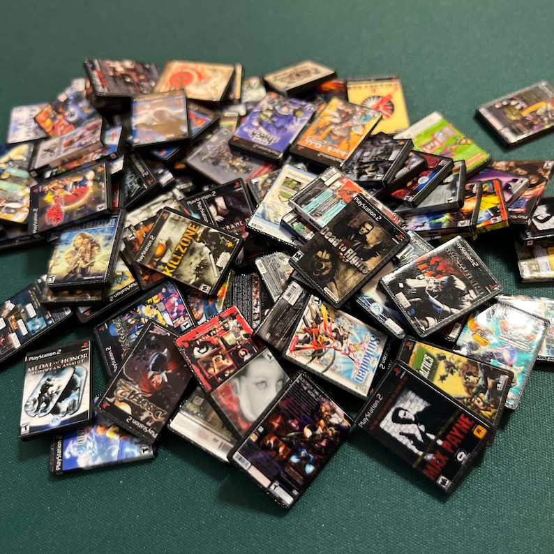 May include: A collection of miniature PlayStation 2 game cases, featuring various game titles. The cases display colorful artwork and game titles, including "Killzone" and "Max Payne". The cases are scattered, creating a visually engaging display.