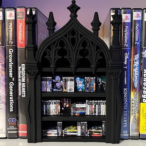 May include: A black gothic-style bookshelf with three shelves, filled with PlayStation 2 video games. The bookshelf has an arched top and decorative detailing. Games include "Kingdom Hearts" and "Yu Yu Hakusho".