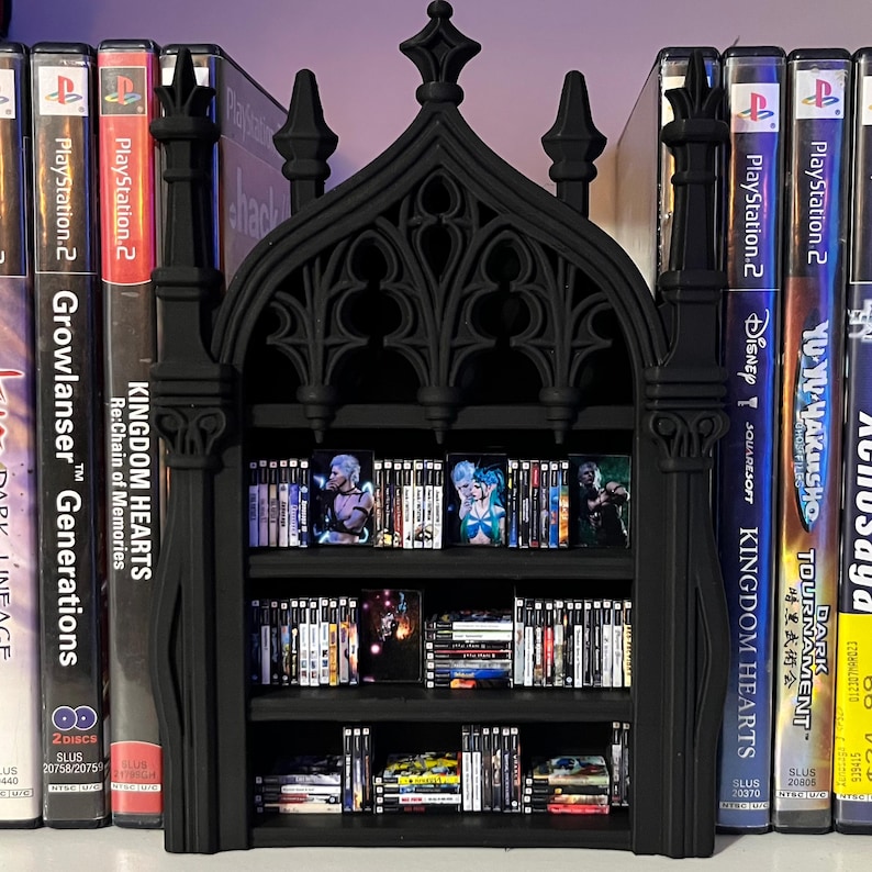 May include: A black gothic-style bookshelf with three shelves, filled with PlayStation 2 video games. The bookshelf has an arched top and decorative detailing. Games include "Kingdom Hearts" and "Yu Yu Hakusho".