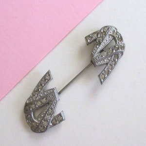 May include: A silver brooch with a pin, featuring a design of two interlocking U shapes decorated with clear rhinestones.