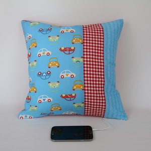 May include: A blue and red pillow with a pattern of cars on one side and a red and white gingham pattern on the other side. The pillow is sitting on a white surface with a black smartphone and white headphones.