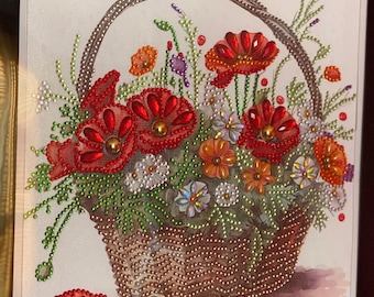 May include: A framed diamond painting of a wicker basket overflowing with red poppies and other colorful flowers. The basket is surrounded by green leaves and the background is white.