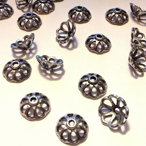 May include: A collection of silver metal bead caps with a decorative, scalloped design. The caps are arranged on a white surface.