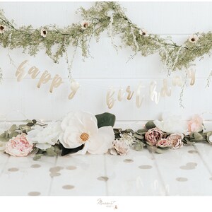 May include: A festive birthday banner with gold lettering that reads "Happy Birthday" hangs above a floral arrangement. The arrangement includes white and pink flowers, greenery, and a garland of small lights.