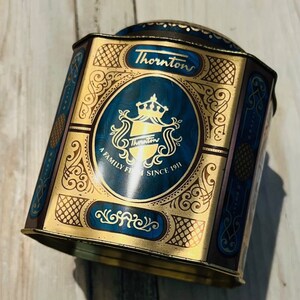 May include: A vintage Thornton's tin with a blue and gold design. The tin features the Thornton's logo, a crown, and the text "A Family Firm Since 1911." The tin has an ornate, decorative pattern and is rectangular with a curved top.