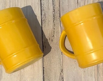 Może przedstawiać: Dwa żółte ceramiczne kubki z uchwytami. Kubki mają cylindryczny kształt z poziomymi paskami wokół korpusu. Kubki są ustawione na jasnej drewnianej powierzchni.