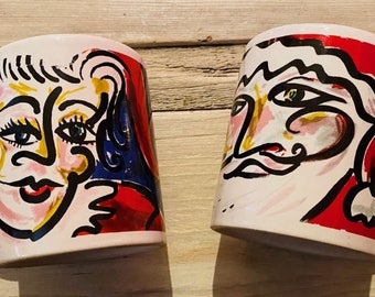 Może przedstawiać: Dwa białe ceramiczne kubki z abstrakcyjnymi wzorami twarzy. Jeden kubek przedstawia twarz kobiety z różowymi, żółtymi, niebieskimi i czarnymi akcentami. Drugi kubek ma twarz mężczyzny z podobnymi kolorami i czerwonym obramowaniem. Oba kubki mają białe uchwyty.