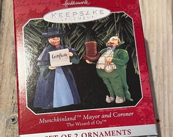 Może przedstawiać: Zestaw ozdób Hallmark Keepsake z dwiema postaciami z Czarnoksiężnika z Oz: burmistrzem i koronerem Munchkinland. Burmistrz trzyma certyfikat, a koroner ma zegarek kieszonkowy. Pudełko jest czerwone z napisem "SET OF 2 ORNAMENTS".