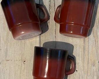 Może przedstawiać: Trzy filiżanki do kawy w stylu vintage o charakterystycznym designie. Kubki mają gradient kolorów, przechodzący od ciemnej czerni u góry do czerwonobrązowego odcienia u podstawy. Każdy kubek ma solidny uchwyt i klasyczny, lekko rozszerzany kształt.