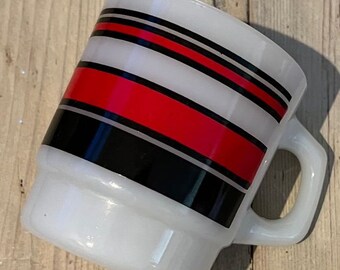 Może przedstawiać: Biały ceramiczny kubek z prostokątnym uchwytem. Kubek posiada poziome pasy czerwonych, czarnych i szarych pasków na białym tle. Kubek jest pochylony, pokazując wzór owijający się wokół korpusu.