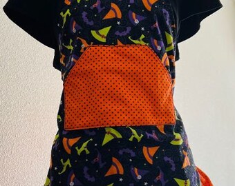 Może przedstawiać: Czarny i pomarańczowy fartuch halloweenowy z nadrukiem czarodziejskiego kapelusza i kieszenią w kropki. Fartuch ma falbankę na dole i idealnie nadaje się do pieczenia lub gotowania.