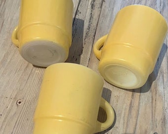 Może przedstawiać: Trzy żółte kubki w stylu vintage z uchwytami. Kubki mają cylindryczny kształt z poziomym paskiem w pobliżu góry i zaokrągloną podstawą. Kubki wykonane są z litego, nieprzezroczystego żółtego materiału.