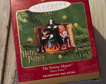 Może przedstawiać: Czerwone pudełko z ozdobą Hallmark Keepsake ze złotymi akcentami, przedstawiające scenę z Harry'ego Pottera. Ozdoba przedstawia postacie wokół kotła. Tekst na pudełku brzmi "The Potions Master" i "DÉCORATION DES FÊTES".