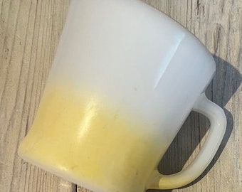 Może przedstawiać: W vintage kubek w kolorze białym i żółtym z efektem ombre, z zakrzywionym uchwytem. Kubek ma gładkie, błyszczące wykończenie i klasyczny, lekko zaokrąglony kształt. Żółty kolor przechodzi w biały, tworząc efekt gradientu. Kubek jest prawdopodobnie wykonany z ceramiki lub szkła.