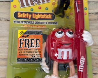 Może przedstawiać: Świecący patyczek bezpieczeństwa M&M's z odłączaną figurką postaci. Opakowanie jest żółte, czerwone i niebieskie. Świecący patyczek jest czerwony i ma logo M&M's. Figurka to czerwona postać M&M's.