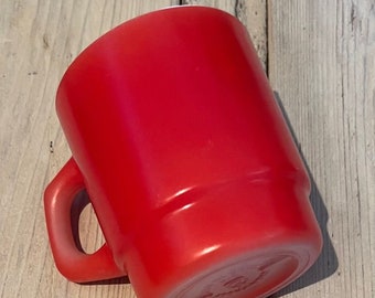 Może przedstawiać: Jasnoczerwony kubek z kwadratowym uchwytem. Kubek wykonany jest z litego, nieprzezroczystego materiału i ma lekko zaokrąglony kształt. Kubek stoi na jasnej drewnianej powierzchni.