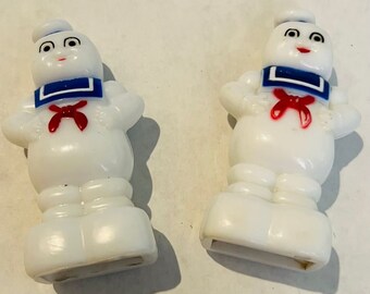 Może przedstawiać: Dwie solniczki i pieprzniczki z białego plastiku w kształcie ludzika Stay Puft Marshmallow Man. Każda figurka ma niebieski kołnierz marynarski, czerwoną chustę i białą czapkę marynarską. Postacie mają czarne oczy, czarne brwi i czerwone usta.