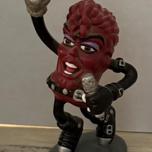 May include: A figurine of a raisin character in a rock star pose. The raisin has a red body, black clothing, and a silver microphone. The raised hand is silver glitter. The figurine stands on a gray oval base.