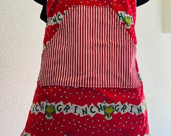 Może przedstawiać: Czerwono-biały pasiasty fartuch z zielonym nadrukiem Grincha i powtarzającymi się napisami "Grinch". Fartuch ma falbankę u dołu i kieszeń z przodu.