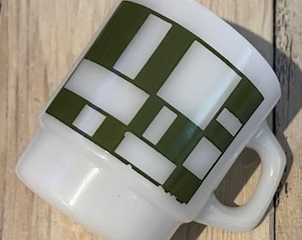 Może przedstawiać: Biały ceramiczny kubek z geometrycznym wzorem w kolorze oliwkowej zieleni. Kubek ma prostokątny uchwyt i lekko rozszerzany brzeg. Wzór składa się z przecinających się prostokątów i kwadratów, tworząc nowoczesny, abstrakcyjny wzór. Kubek stoi na drewnianej powierzchni.