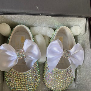Puede incluir: Un par de zapatos de beb&eacute; blancos con un lazo de sat&eacute;n blanco y pedrer&iacute;a iridiscente.