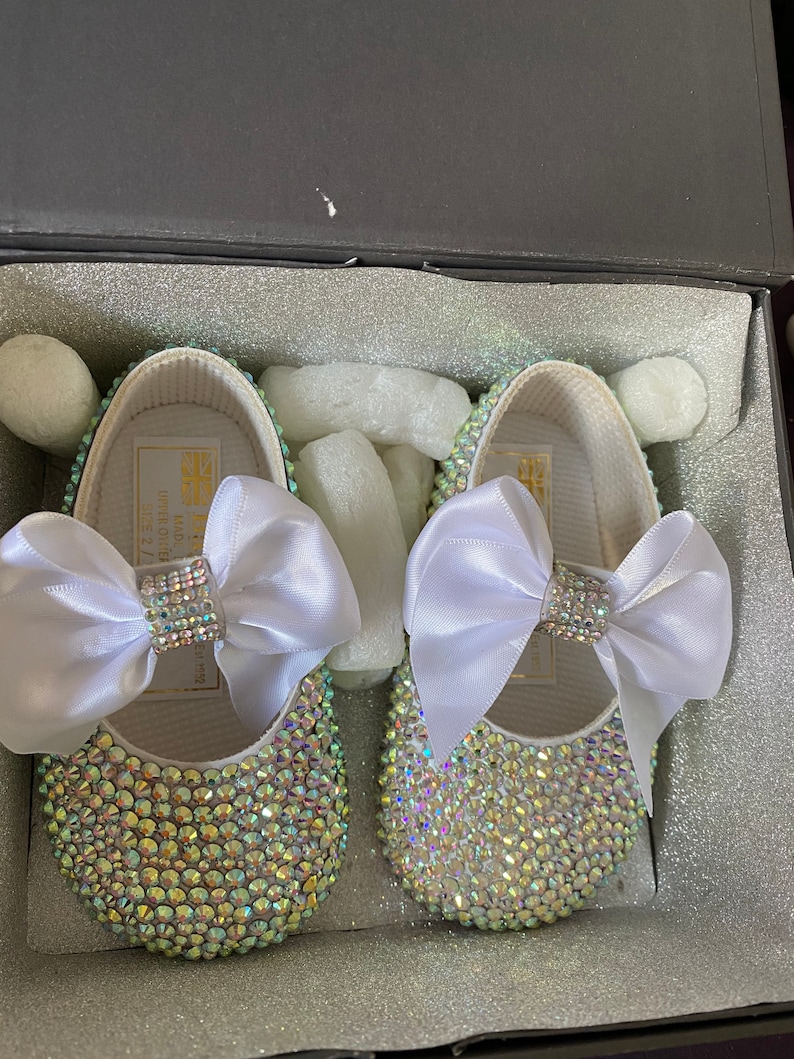 Puede incluir: Un par de zapatos de beb&eacute; blancos con un lazo de sat&eacute;n blanco y pedrer&iacute;a iridiscente.