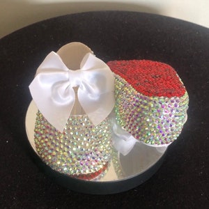 Puede incluir: Un par de zapatos de beb&eacute; decorados con pedrer&iacute;a iridiscente y un lazo de sat&eacute;n blanco. Un zapato tiene pedrer&iacute;a roja en la punta.