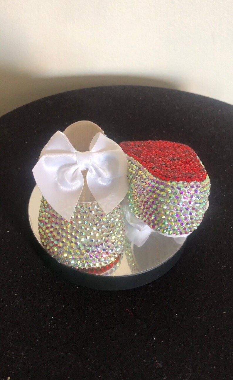 Puede incluir: Un par de zapatos de beb&eacute; decorados con pedrer&iacute;a iridiscente y un lazo de sat&eacute;n blanco. Un zapato tiene pedrer&iacute;a roja en la punta.