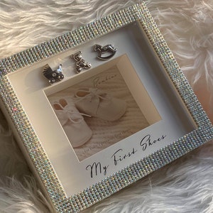 Puede incluir: Marco de fotos blanco con un borde brillante. El marco tiene una foto de zapatos de beb&eacute; y el texto "My First Shoes" y "for baby's precious first shoes". Tambi&eacute;n hay peque&ntilde;os amuletos plateados de un caballo de balanc&iacute;n, un osito de peluche y un cochecito.