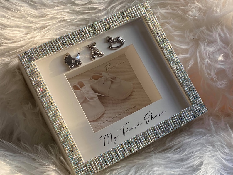 Puede incluir: Marco de fotos blanco con un borde brillante. El marco tiene una foto de zapatos de beb&eacute; y el texto "My First Shoes" y "for baby's precious first shoes". Tambi&eacute;n hay peque&ntilde;os amuletos plateados de un caballo de balanc&iacute;n, un osito de peluche y un cochecito.