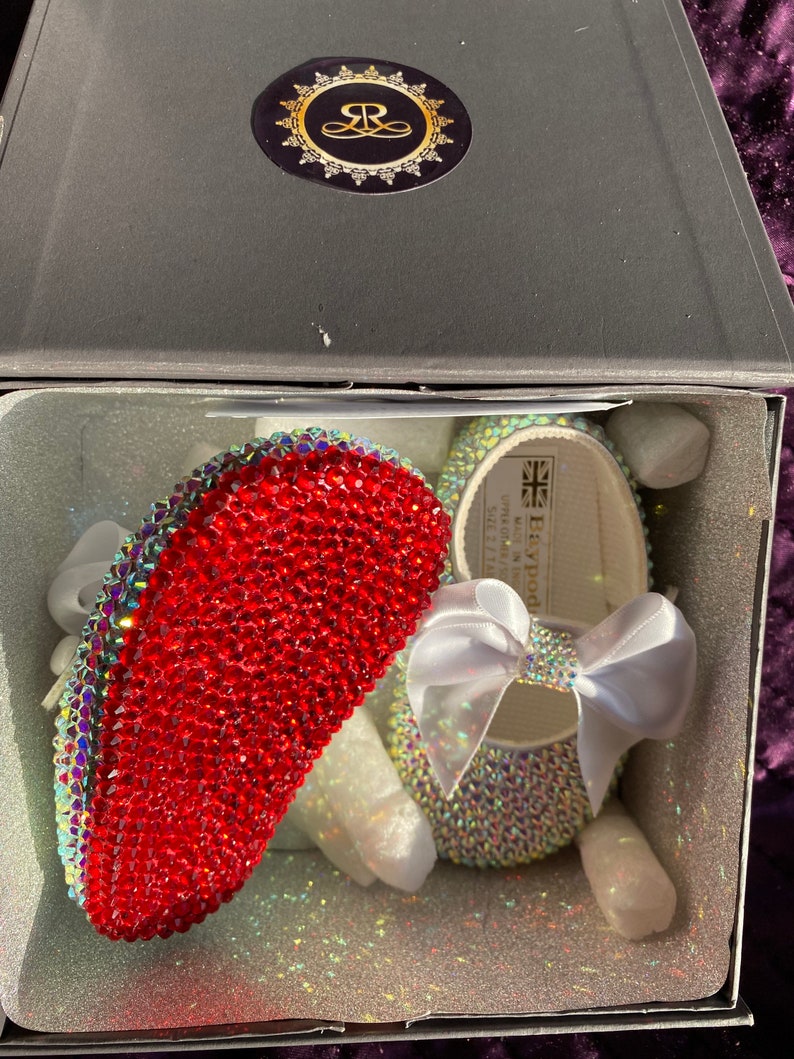 Puede incluir: Un par de zapatos de beb&eacute;, uno rojo y otro blanco, cubiertos de brillantes de cristal. Los zapatos est&aacute;n en una caja blanca con un logotipo dorado que dice "Heywood".
