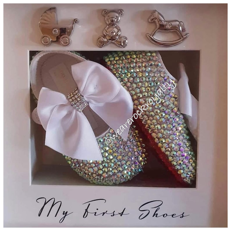 Puede incluir: Un marco blanco con un par de zapatos de beb&eacute; decorados con pedrer&iacute;a iridiscente. Los zapatos tienen un lazo de sat&eacute;n blanco y el texto "My First Shoes" est&aacute; impreso debajo de los zapatos.