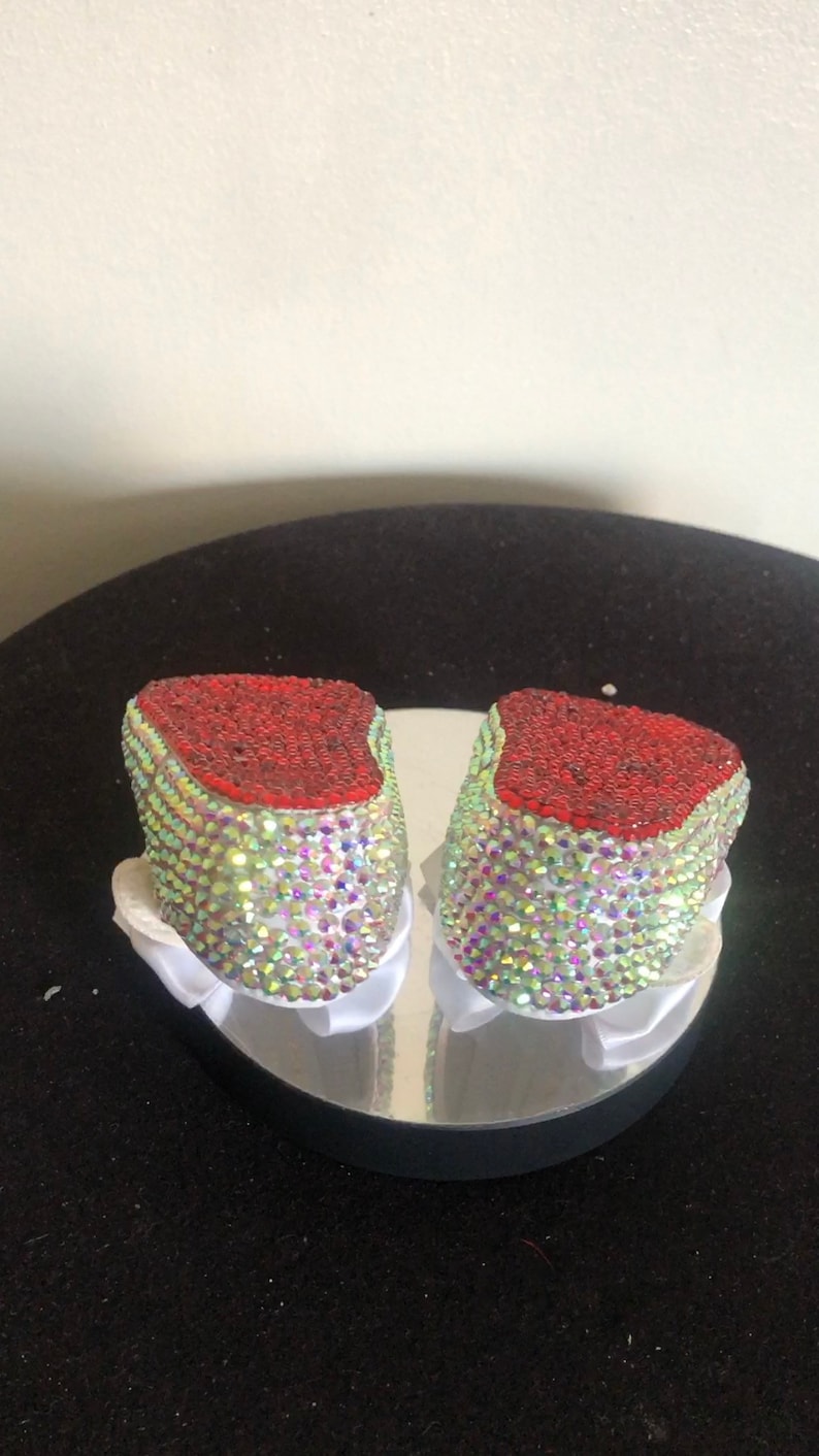 Puede incluir: Un par de zapatos de beb&eacute; decorados con pedrer&iacute;a iridiscente y ribete rojo.