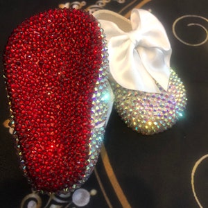 Puede incluir: Un par de zapatos de beb&eacute; con un lazo de sat&eacute;n blanco. Un zapato est&aacute; cubierto de pedrer&iacute;a roja y el otro de pedrer&iacute;a iridiscente.
