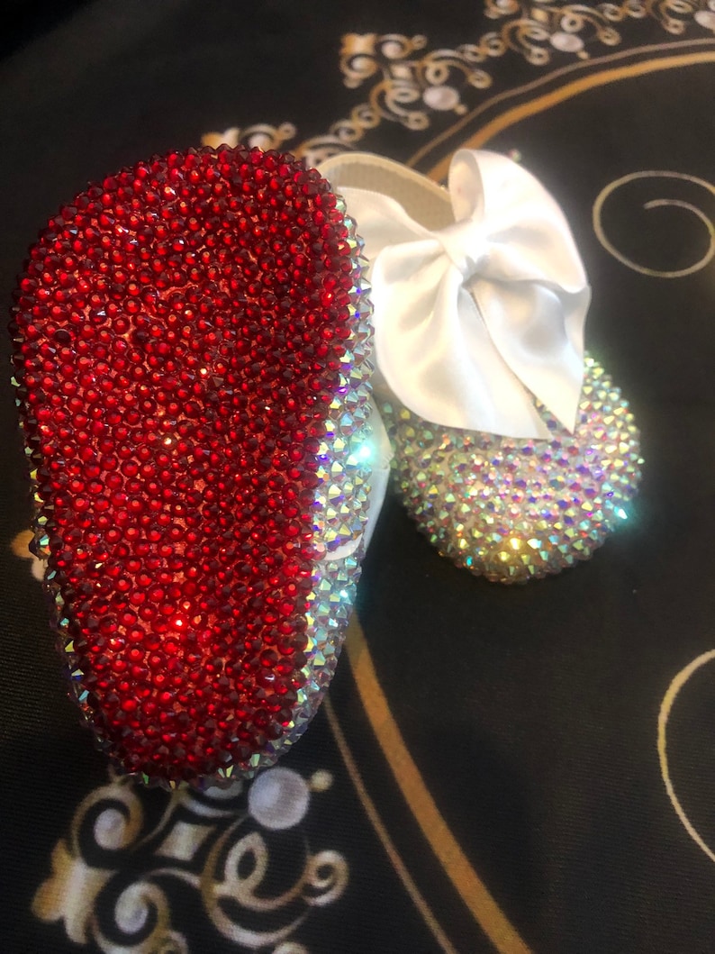 Puede incluir: Un par de zapatos de beb&eacute; con un lazo de sat&eacute;n blanco. Un zapato est&aacute; cubierto de pedrer&iacute;a roja y el otro de pedrer&iacute;a iridiscente.