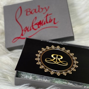 Puede incluir: Una caja negra con un logotipo dorado que presenta una "R" estilizada dentro de un c&iacute;rculo con un borde de diamantes. La caja est&aacute; abierta y el interior est&aacute; forrado con papel de seda blanco. La caja est&aacute; sentada sobre una superficie blanca y esponjosa. La parte superior de la caja es gris y tiene el texto "Baby Louboutin" en letras rojas.