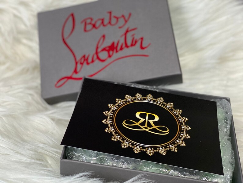 Puede incluir: Una caja negra con un logotipo dorado que presenta una "R" estilizada dentro de un c&iacute;rculo con un borde de diamantes. La caja est&aacute; abierta y el interior est&aacute; forrado con papel de seda blanco. La caja est&aacute; sentada sobre una superficie blanca y esponjosa. La parte superior de la caja es gris y tiene el texto "Baby Louboutin" en letras rojas.