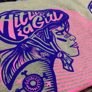 K&ouml;nnte beinhalten: Ein graues T-Shirt mit einem pinkfarbenen und blauen Grafikdesign. Die Grafik zeigt eine Frau, die einen Helm tr&auml;gt, mit dem Text "Hit Like a Girl" &uuml;ber ihrem Kopf. Die Grafik enth&auml;lt auch ein pinkfarbenes und blaues Streifenmuster.