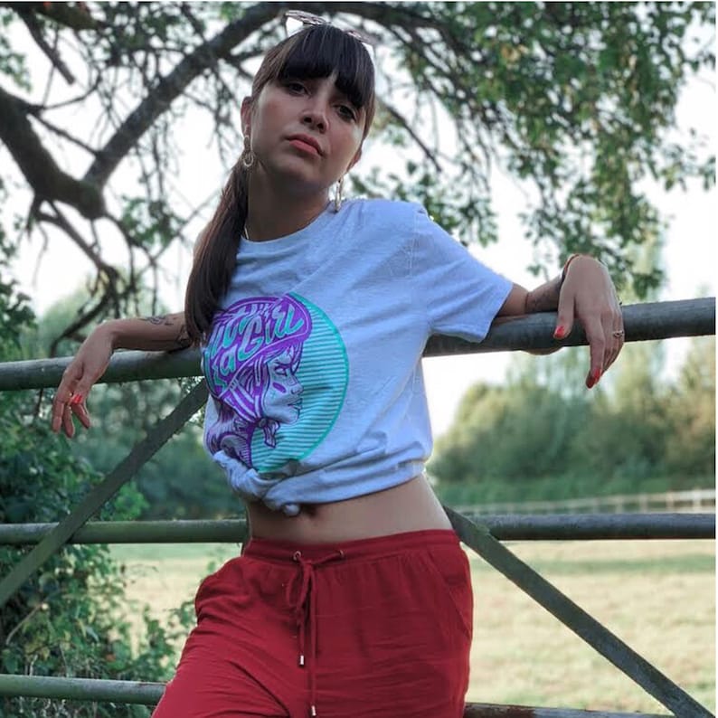 K&ouml;nnte beinhalten: Ein wei&szlig;es T-Shirt mit einem lila und gr&uuml;nen Grafikdruck eines Frauen Gesichts mit dem Text "Bad Girl" darauf. Die Frau tr&auml;gt ein lila Stirnband und hat einen gr&uuml;n-wei&szlig; gestreiften Hintergrund.