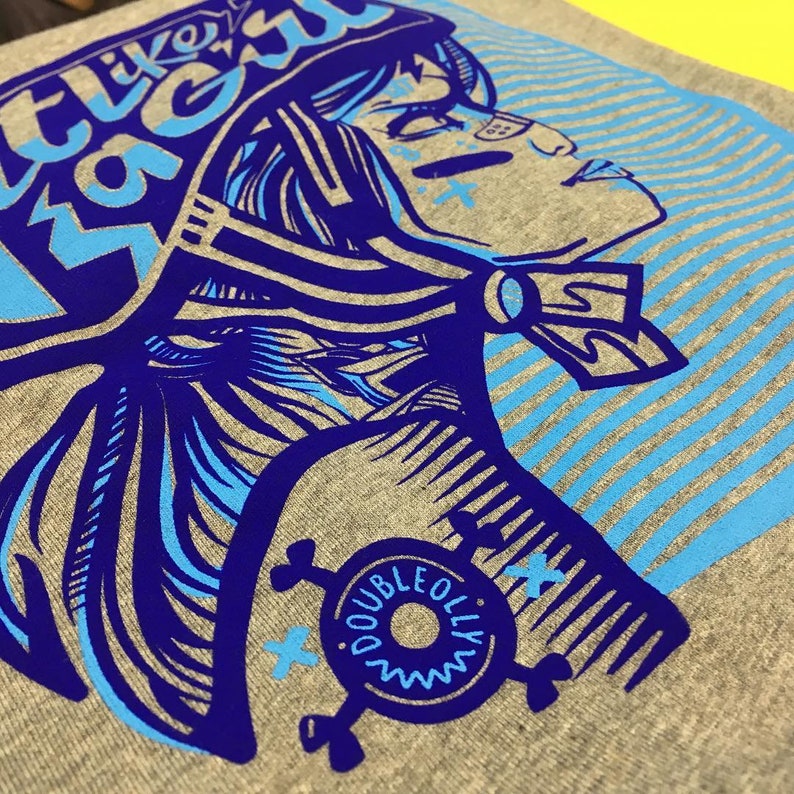 K&ouml;nnte beinhalten: Ein graues T-Shirt mit einem blauen und wei&szlig;en Grafikdruck einer Frau mit Helm. Die Grafik enth&auml;lt den Text "Feel Like I'm Flying" und ein kreisf&ouml;rmiges Logo mit dem Text "Double Olly".