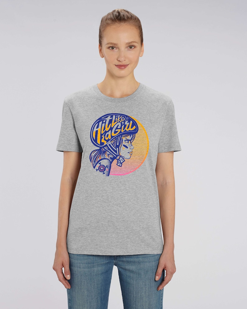 K&ouml;nnte beinhalten: Graues T-Shirt mit einem Grafikdruck einer Frau mit Helm und dem Text "Hit Like a Girl" in einem blau-orangenen Farbschema.