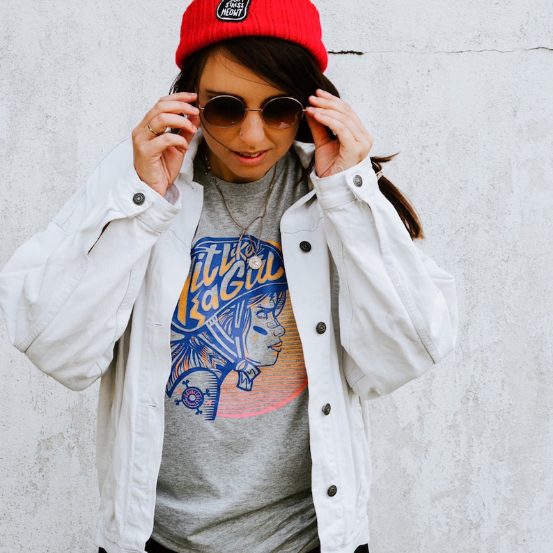 K&ouml;nnte beinhalten: Eine Frau tr&auml;gt eine wei&szlig;e Jeansjacke &uuml;ber einem grauen T-Shirt mit einem Grafikdruck einer Frau mit Helm und dem Text "Hit Like a Girl". Sie tr&auml;gt au&szlig;erdem eine rote M&uuml;tze und eine runde Sonnenbrille.