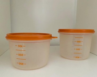 Puede incluir: Dos recipientes de plástico transparente con tapas naranjas. Los recipientes tienen marcas de medición en mililitros.