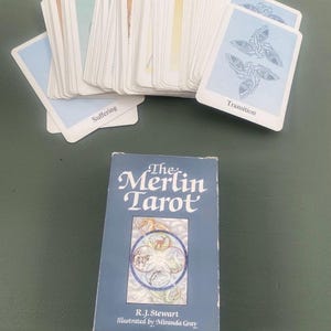 May include: A deck of The Merlin Tarot cards by R.J. Stewart and illustrated by Miranda Gray, featuring a blue box with a circular animal illustration. The cards are fanned out, displaying light blue backs with a knotwork design and the text Transition.